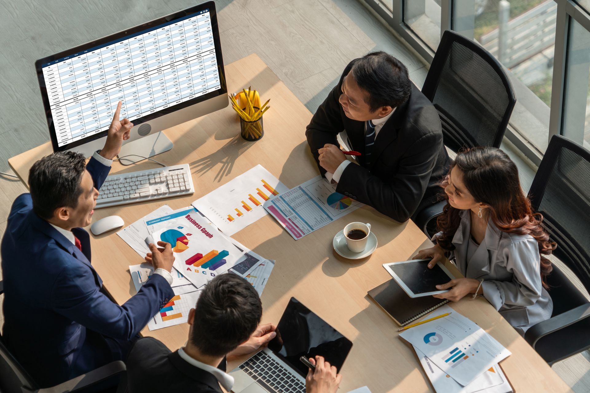 Business team reviewing data and charts around a conference table. One points at a computer screen.