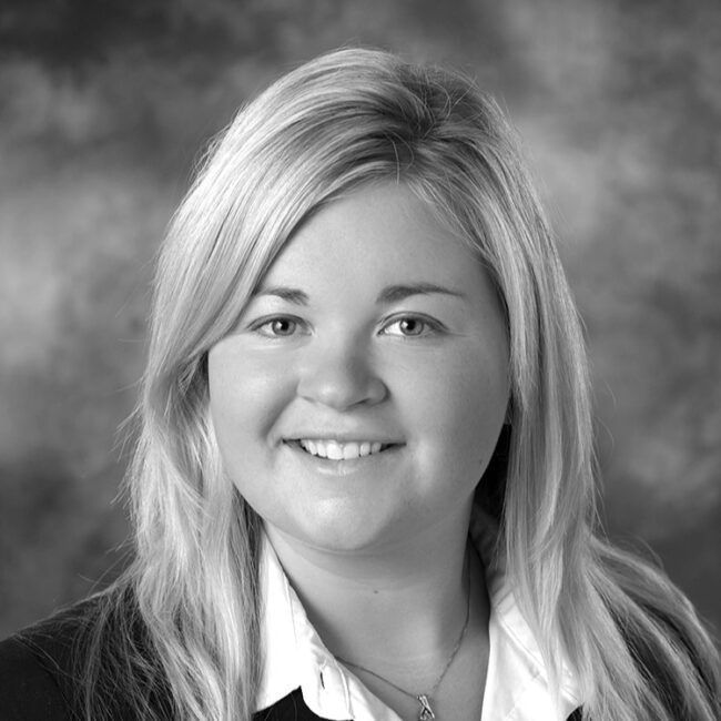 Blonde woman smiling, wearing a suit jacket and white collared shirt, against a blurred background.