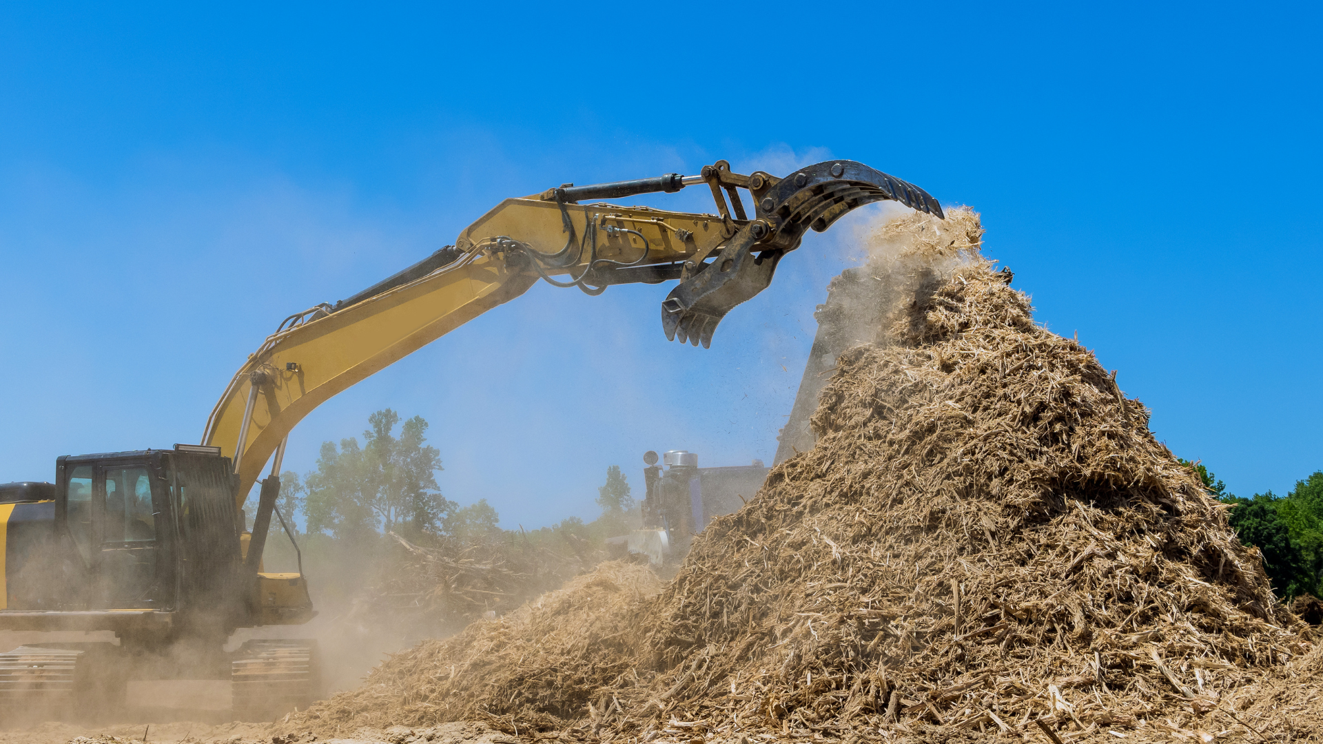 Excavator loading a large woodchip pile at a construction or recycling site under a blue sky