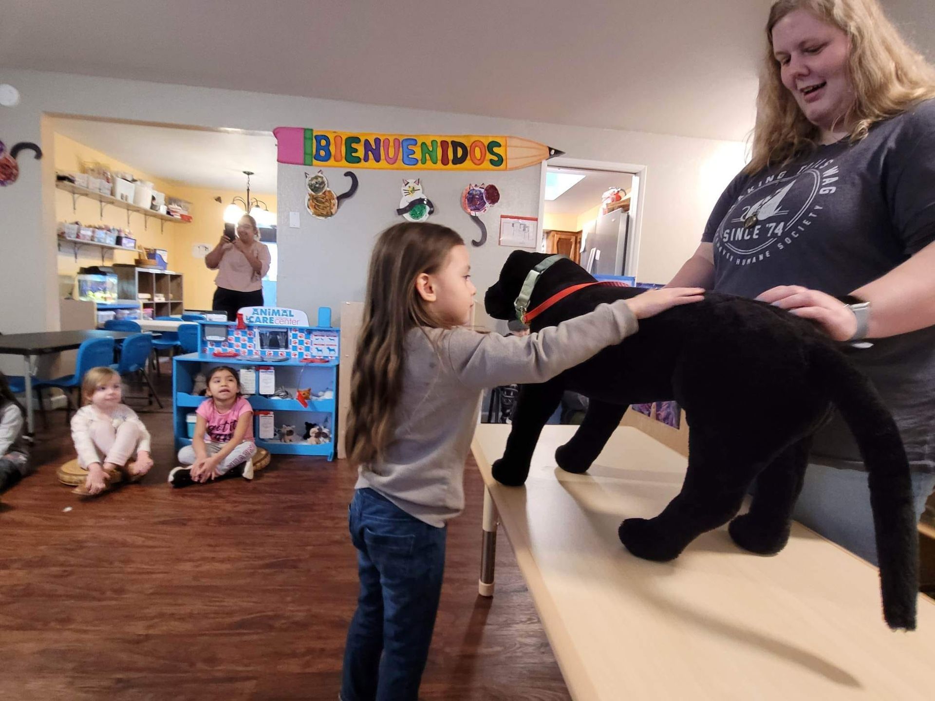 A little girl is petting a stuffed animal in front of a sign that says bienvenidos