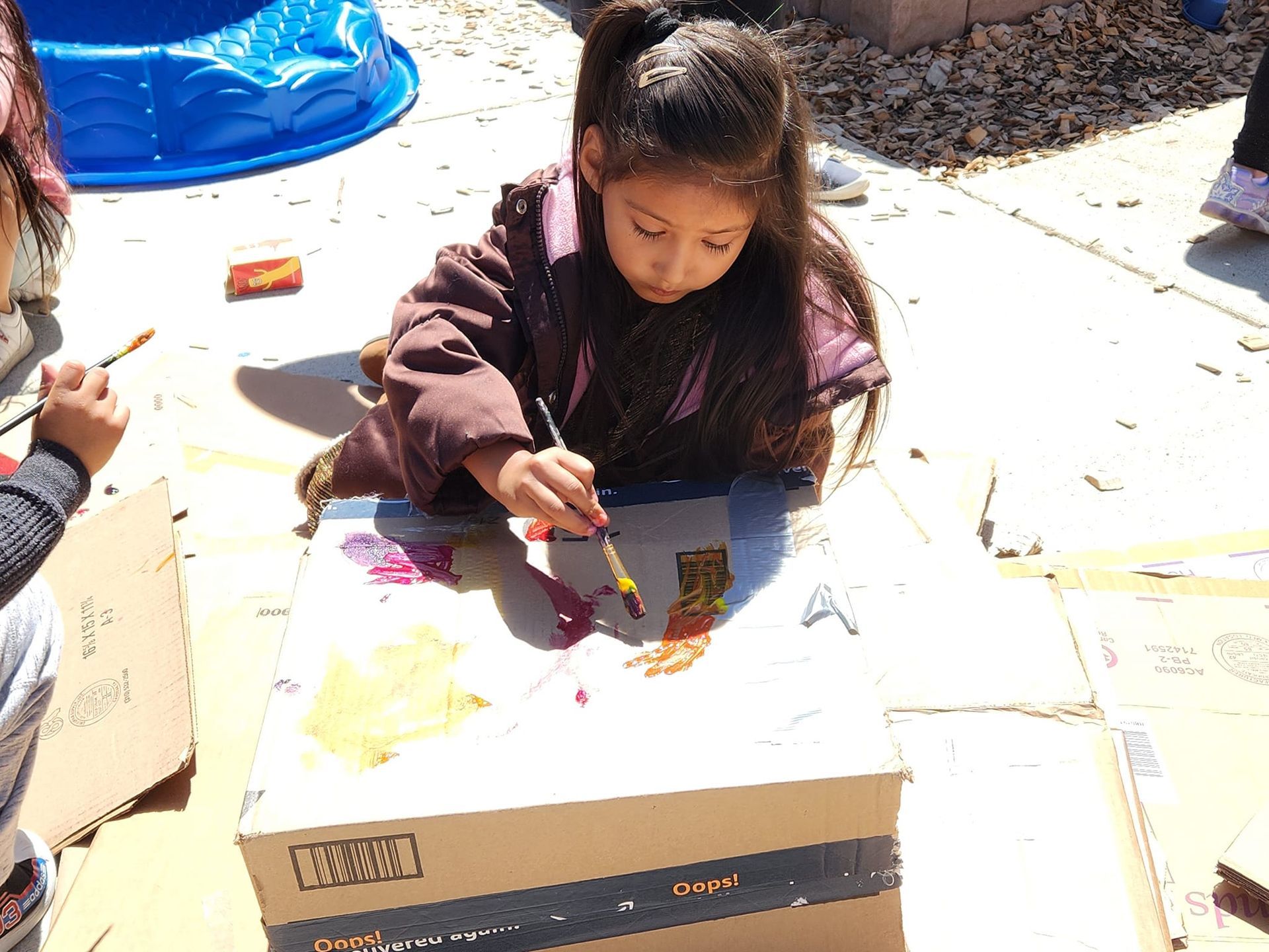 A little girl is painting on a cardboard box with a brush.