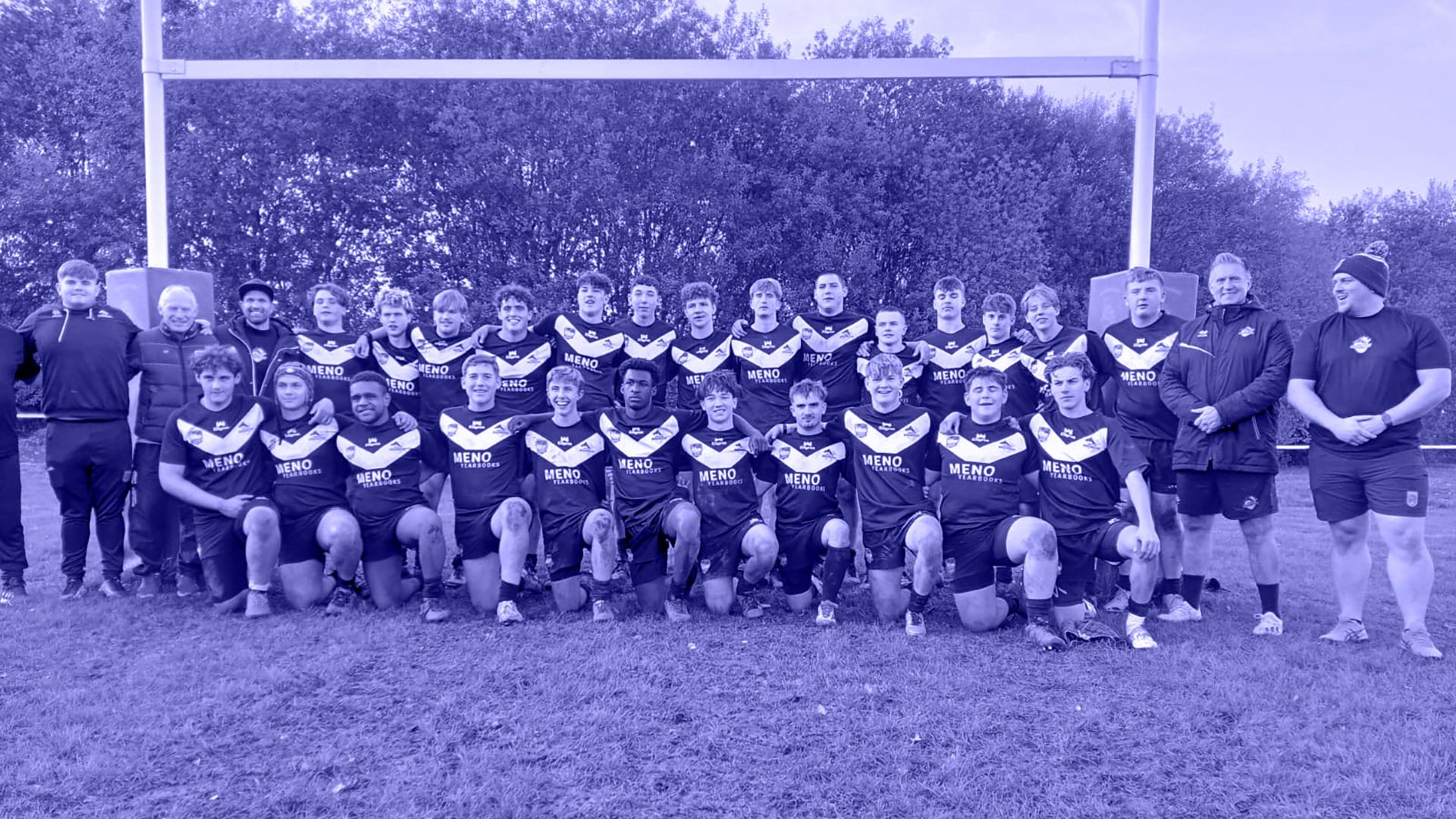 Rugby team posing on a field. Dark uniforms, goal posts behind them.