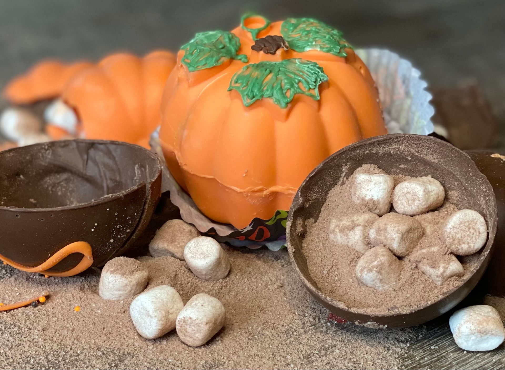 Fall-themed display with orange pumpkins on hot cocoa beside chocolate shells and stones. Hot Co Explosions cocoa bombs.