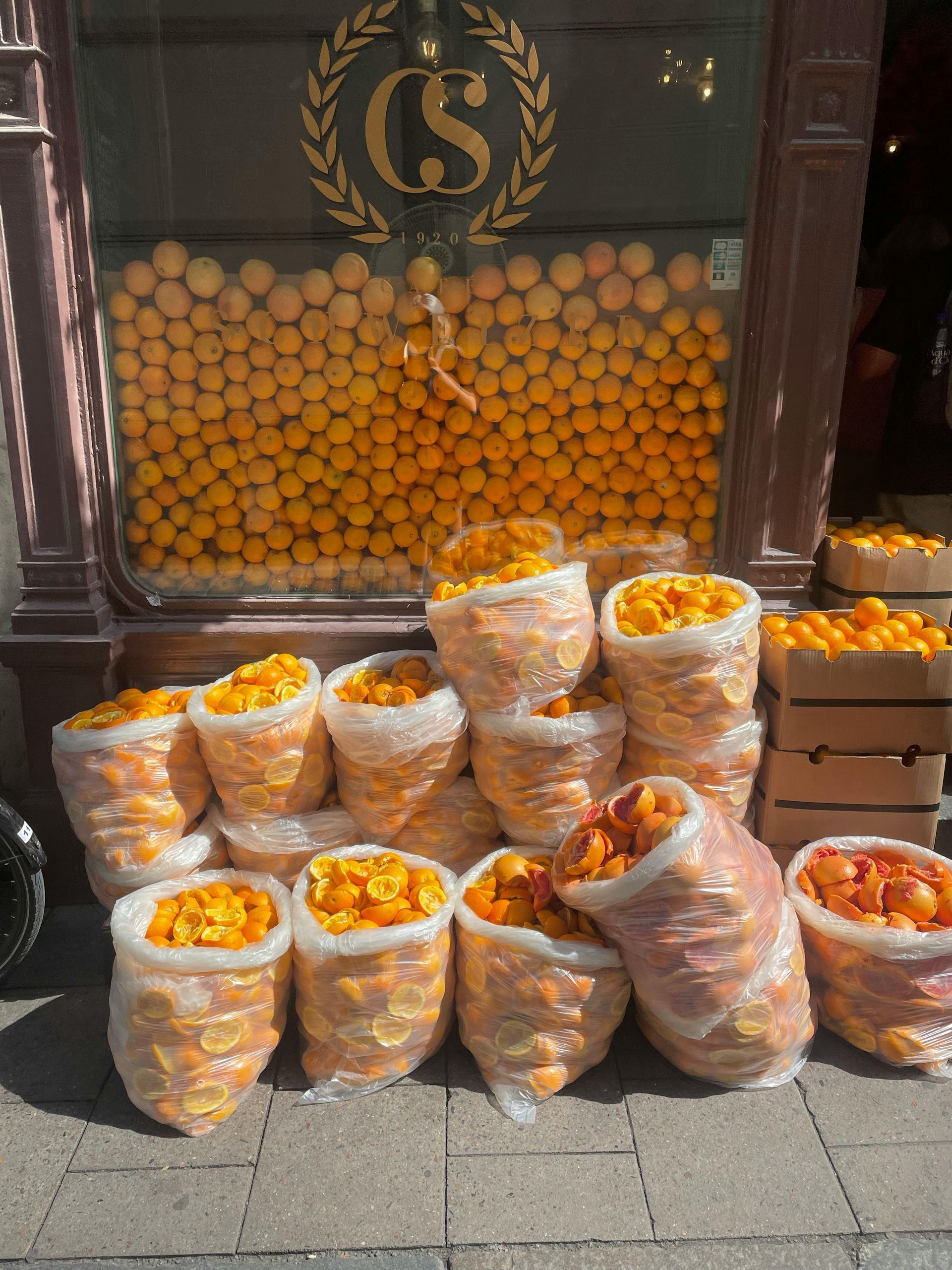 Stacks of clear bags filled with oranges outside a shop, with more oranges displayed behind a window.
