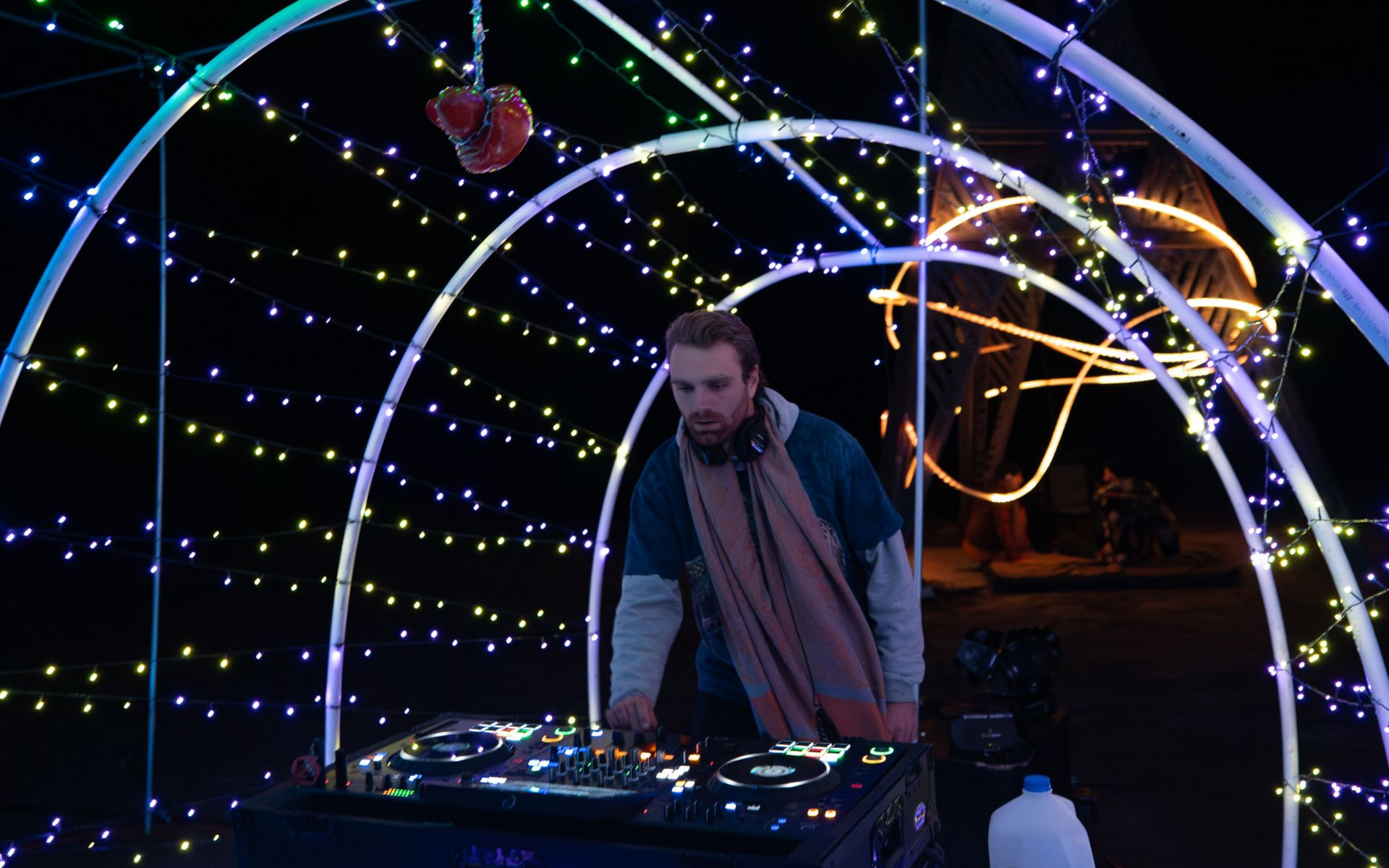 DJ mixing music under illuminated archway, wearing a scarf.