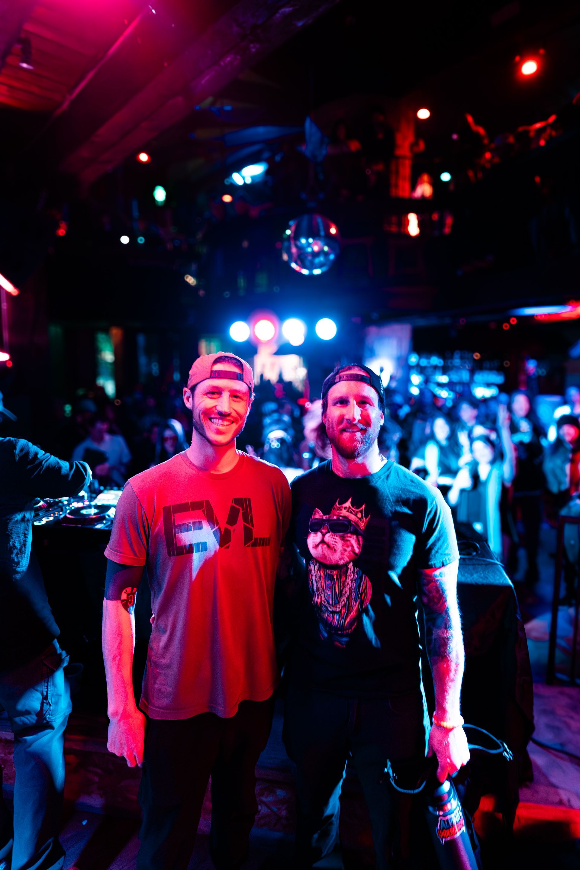 Two men, wearing hats, standing in a crowded, dimly lit club, under a disco ball. One man has tattoos and holds a drink.