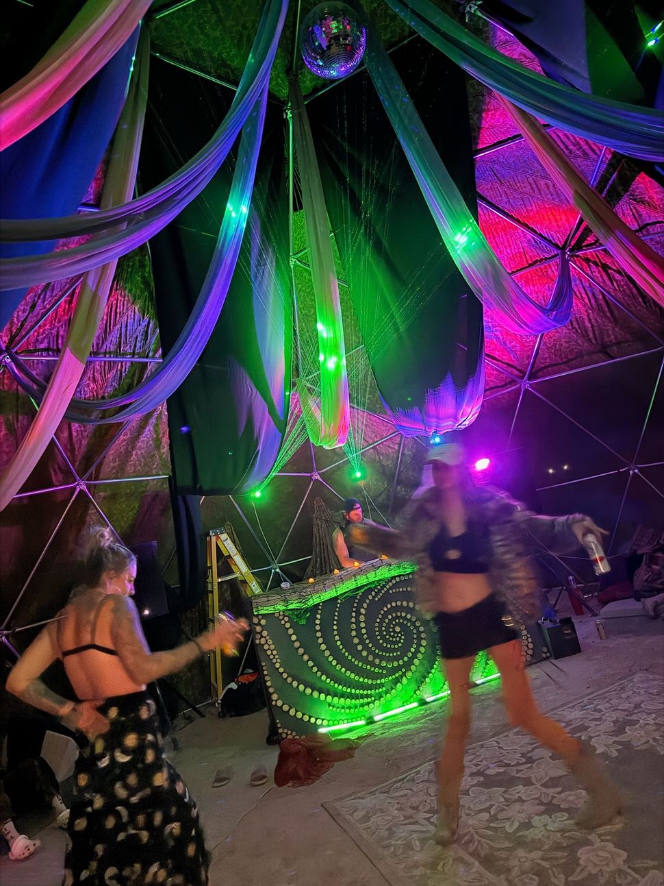 Dancers perform under neon-lit fabric canopies, with glowing green and purple lights in a festive indoor setting