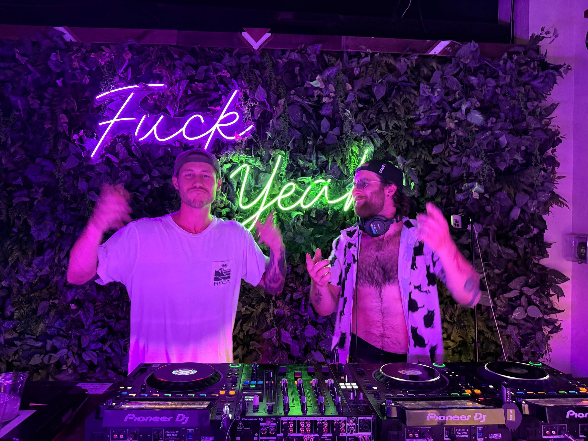 Two DJs at a party, illuminated by neon sign