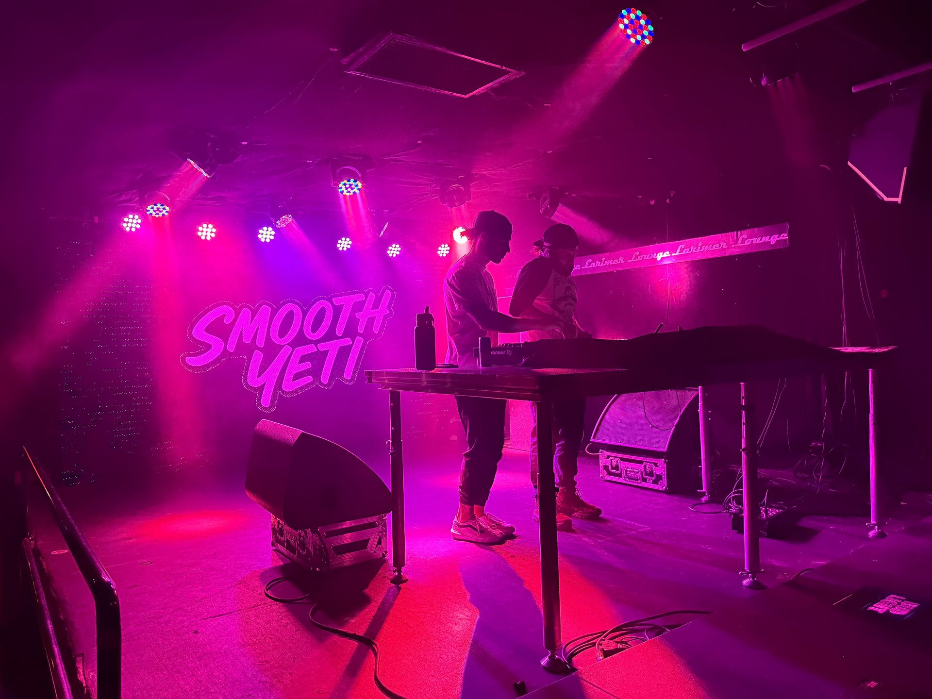 Two figures perform at a music venue. Pink lighting bathes the stage.