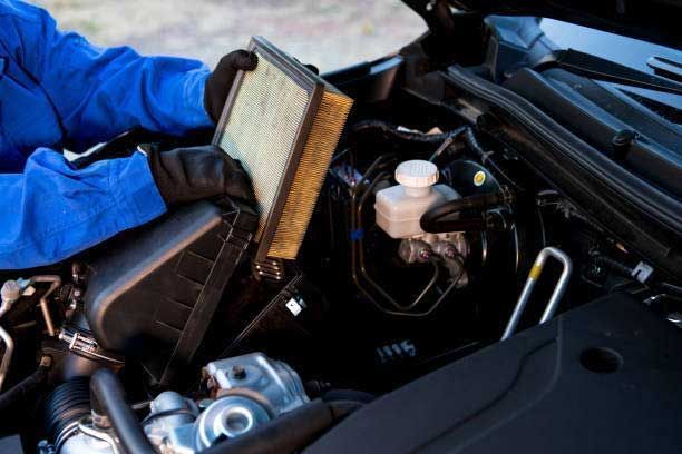 Mechanic wearing blue gloves holds a car air filter over an open engine bay with tools nearby