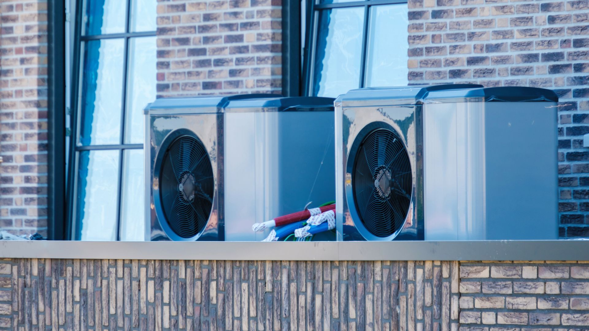 Two outdoor HVAC units on a brick ledge beside a windowed brick wall Two outdoor HVAC units on a brick ledge beside a windowed brick wall