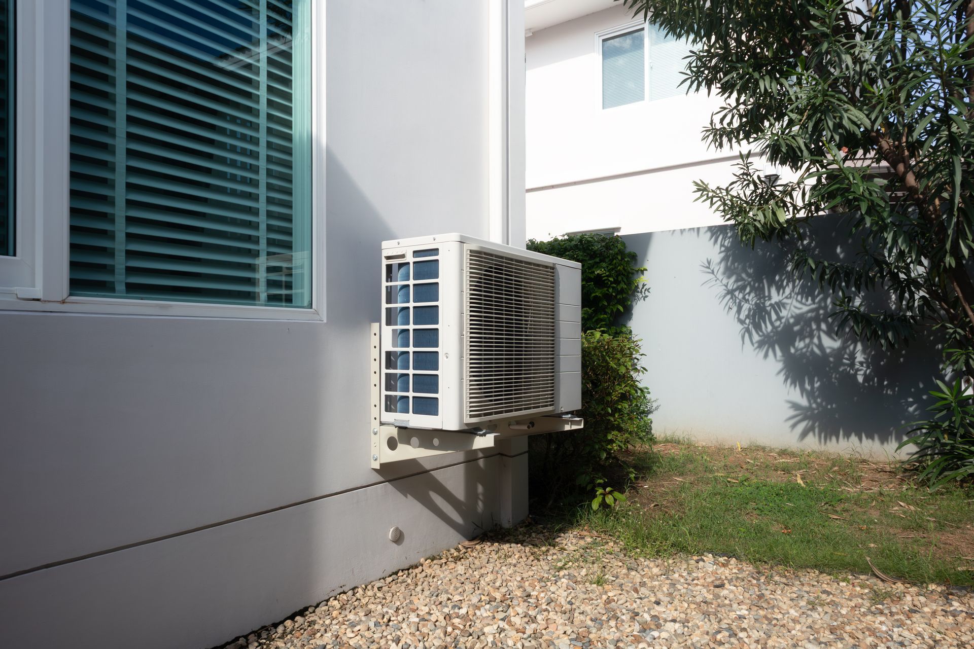 Outdoor air-conditioning unit beside a white house wall and window, next to a grassy yard. Outdoor air-conditioning unit beside a white house wall and window, next to a grassy yard.