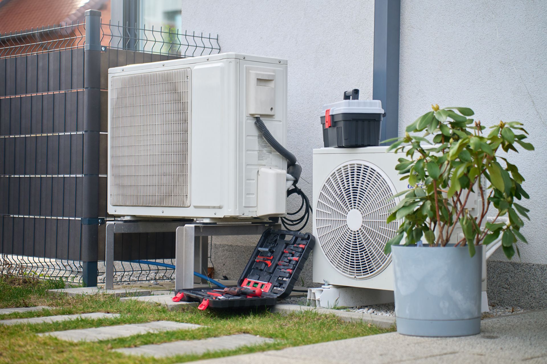Outdoor air conditioner units beside a gate, with tools on the ground and a potted plant nearby. Outdoor air conditioner units beside a gate, with tools on the ground and a potted plant nearby.