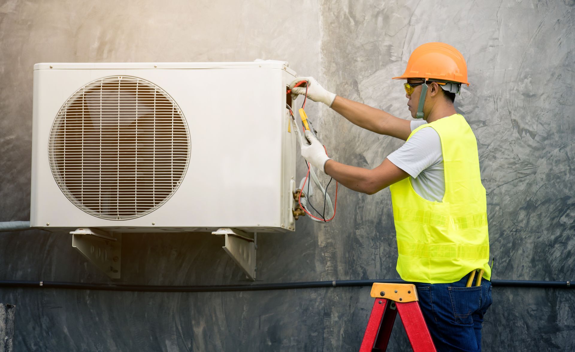 Technician in orange helmet and yellow vest repairs an outdoor air conditioner unit. Technician in orange helmet and yellow vest repairs an outdoor air conditioner unit.