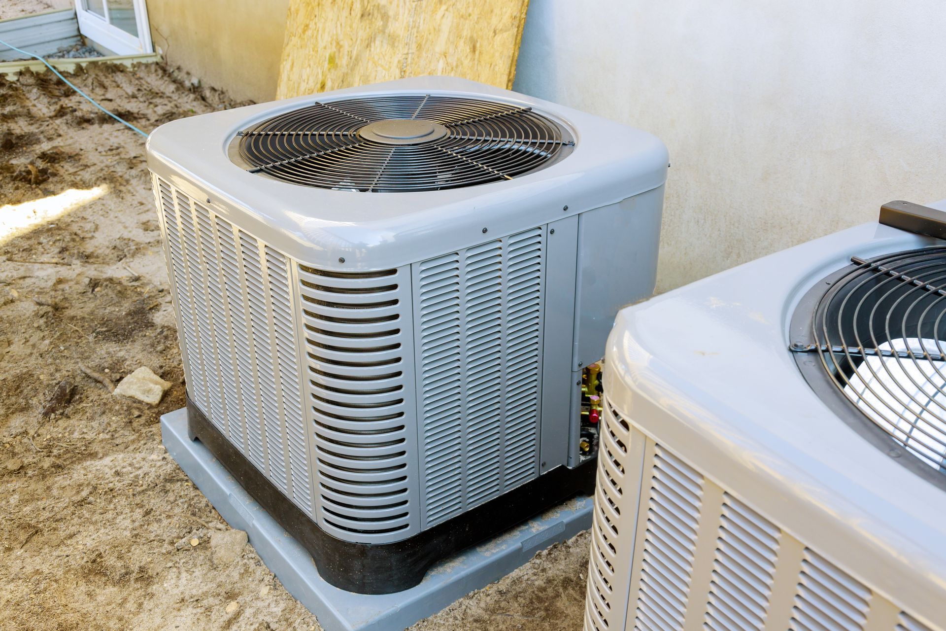 Two gray outdoor air conditioning units beside a building on sandy ground Two gray outdoor air conditioning units beside a building on sandy ground