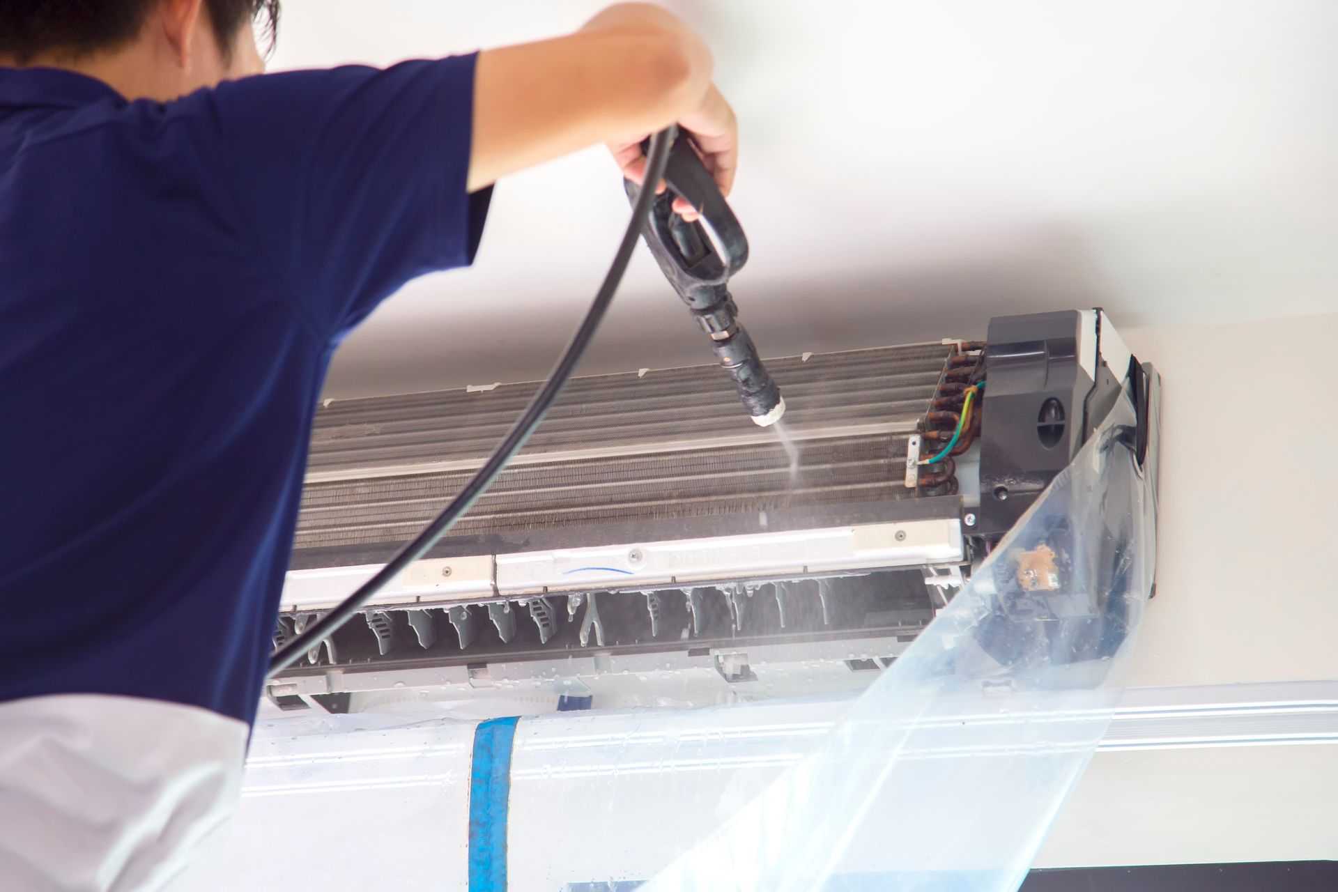 Technician cleaning a wall-mounted air conditioner with a spray hose and protective plastic cover Technician cleaning a wall-mounted air conditioner with a spray hose and protective plastic cover