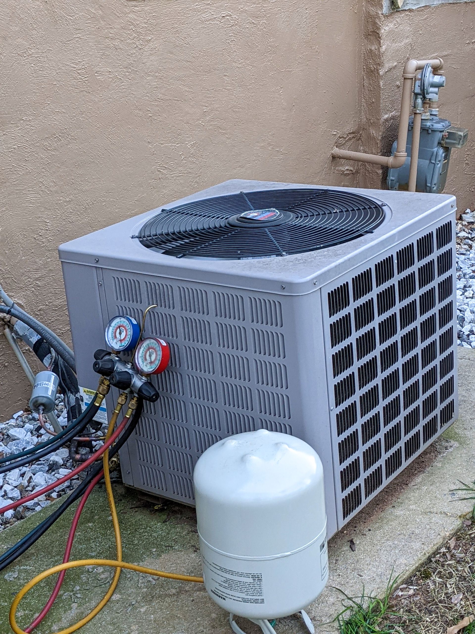 Outdoor air-conditioning unit with gauges and hoses attached, beside a white filter canister against a wall Outdoor air-conditioning unit with gauges and hoses attached, beside a white filter canister against a wall