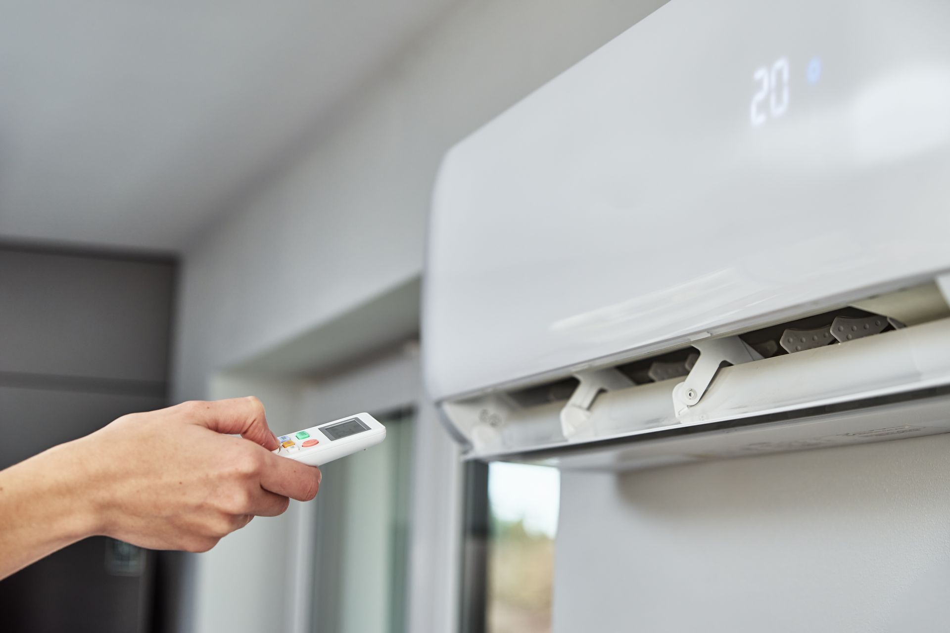 Hand holding a remote pointed at a wall-mounted air conditioner with the display on. Hand holding a remote pointed at a wall-mounted air conditioner with the display on.