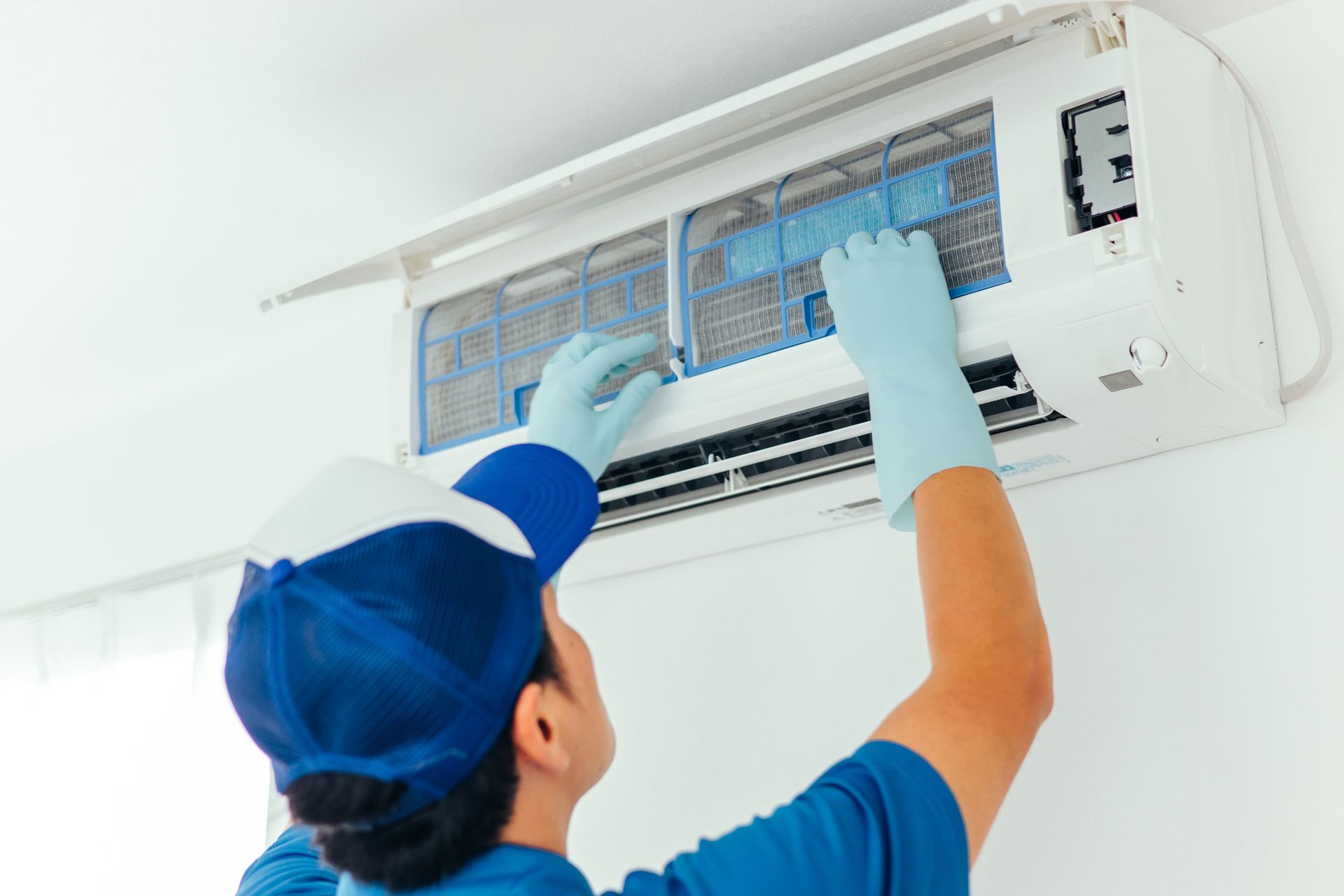 Technician cleaning an air conditioner filter on a wall-mounted unit with gloves and a blue cap Technician cleaning an air conditioner filter on a wall-mounted unit with gloves and a blue cap
