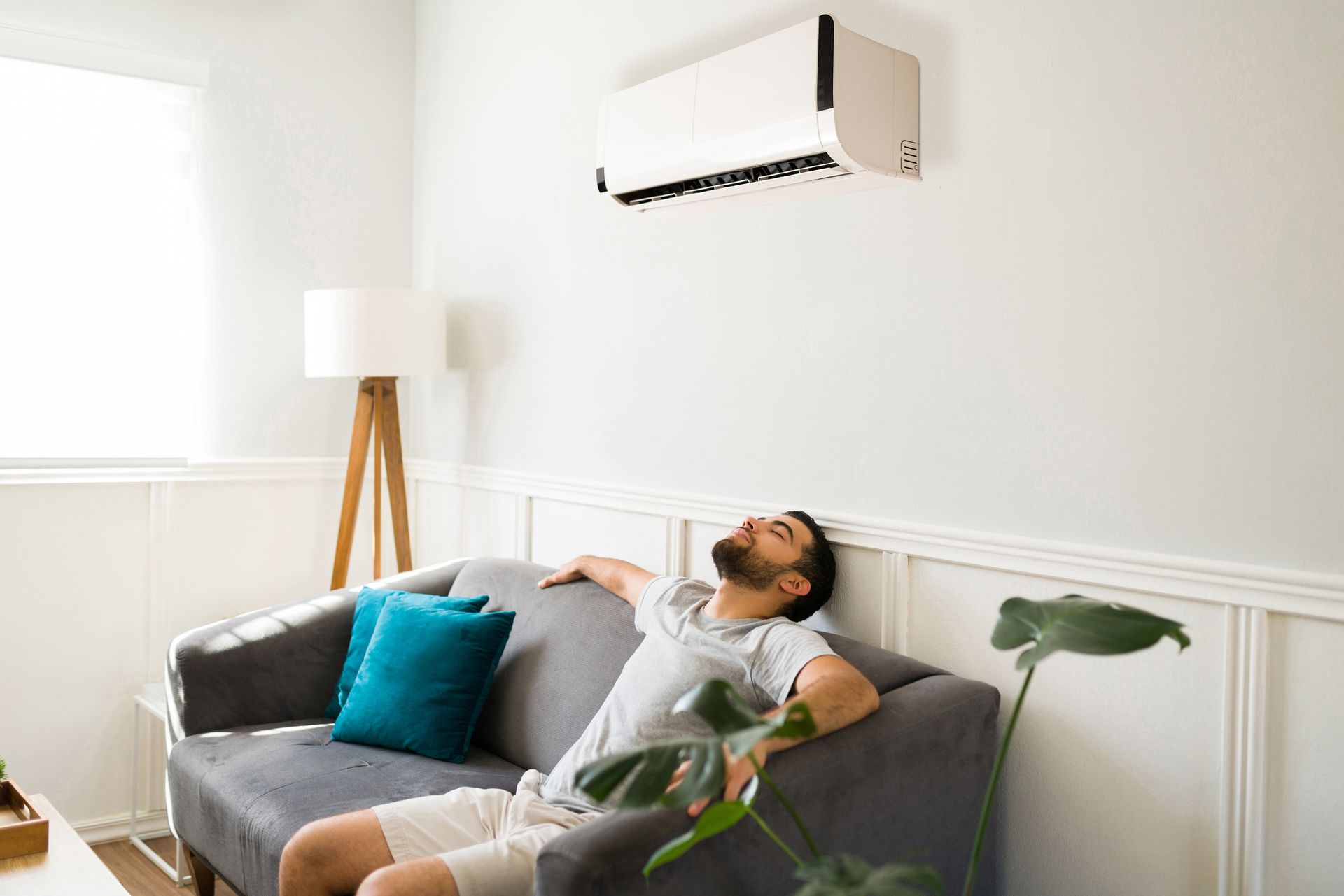 Man reclining on a gray sofa beneath a wall-mounted air conditioner in a bright living room Man reclining on a gray sofa beneath a wall-mounted air conditioner in a bright living room