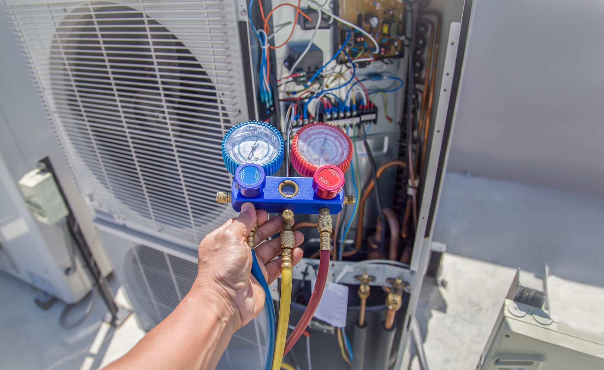 Hand holding a blue-red gauge manifold on an outdoor HVAC unit with exposed wiring and pipes Hand holding a blue-red gauge manifold on an outdoor HVAC unit with exposed wiring and pipes