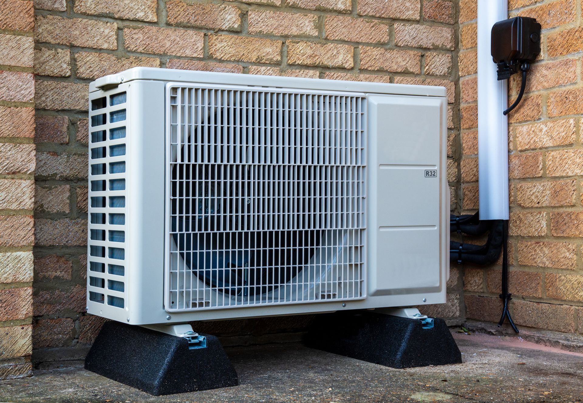 Outdoor air conditioning unit on black stands beside a brick wall and downspout. Outdoor air conditioning unit on black stands beside a brick wall and downspout.