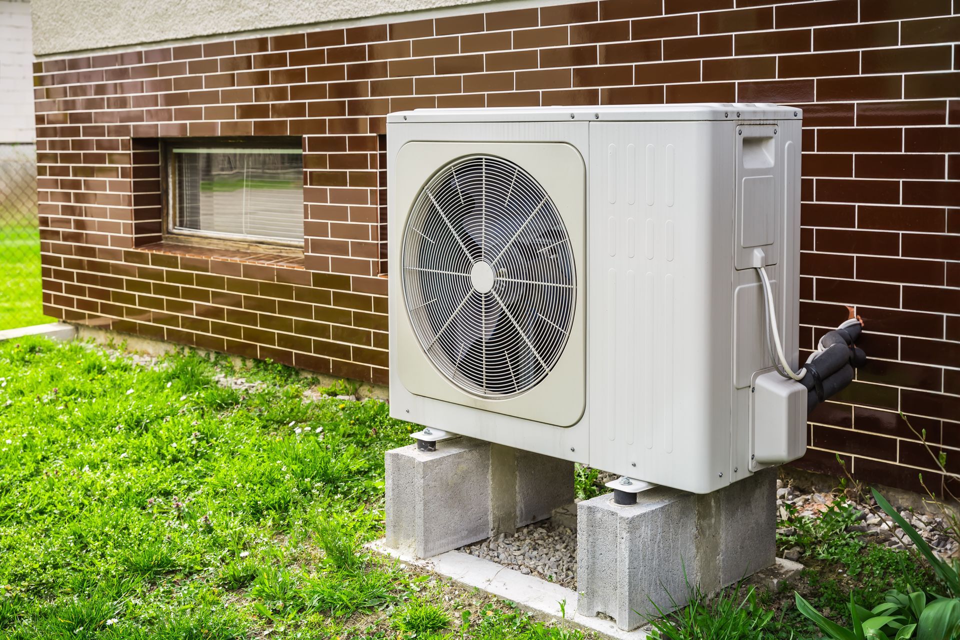 Outdoor air conditioner unit beside a brick wall and window on cinder blocks in a grassy yard Outdoor air conditioner unit beside a brick wall and window on cinder blocks in a grassy yard