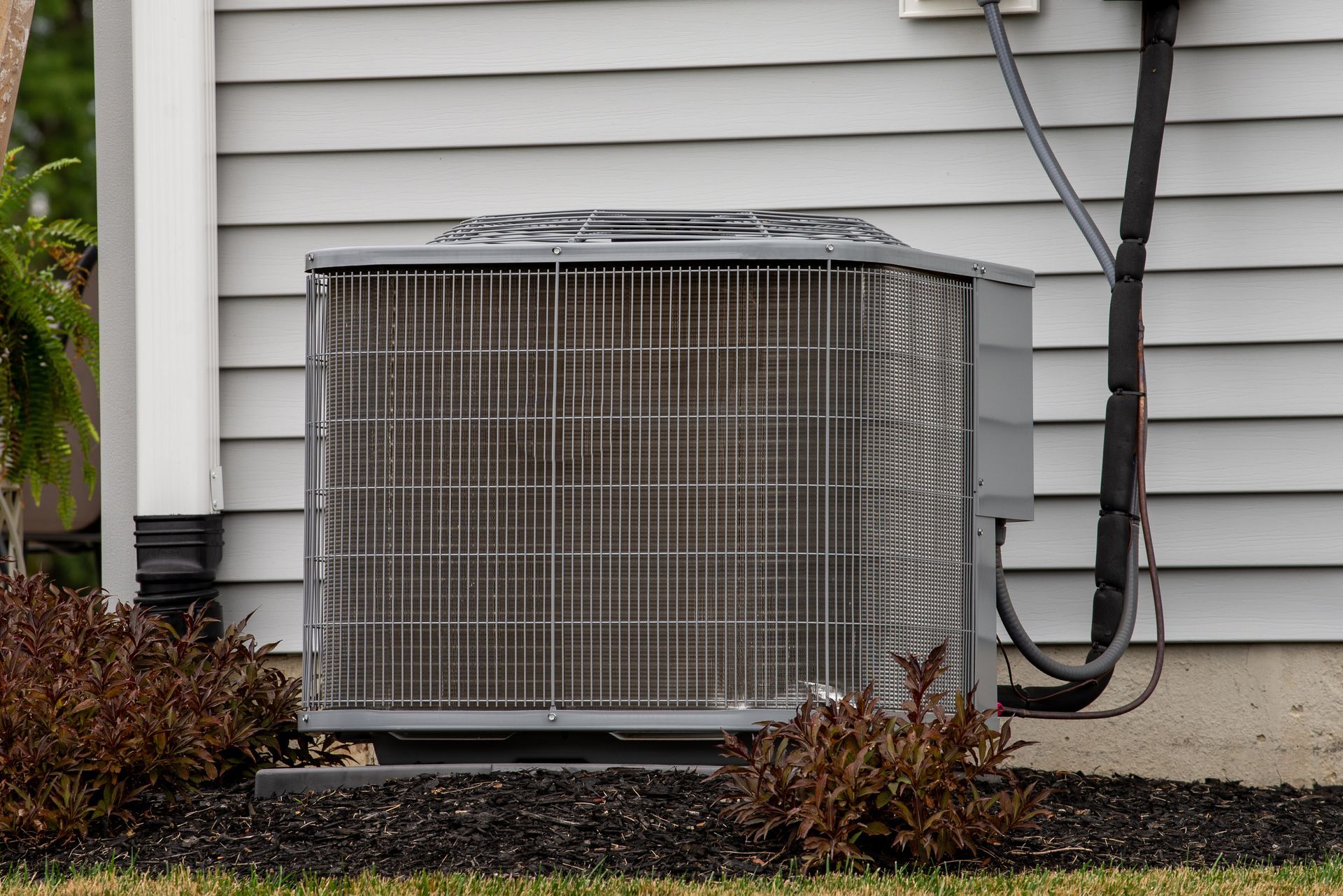 Outdoor air conditioning unit beside a house wall, with shrubs and a coiled hose nearby. Outdoor air conditioning unit beside a house wall, with shrubs and a coiled hose nearby.