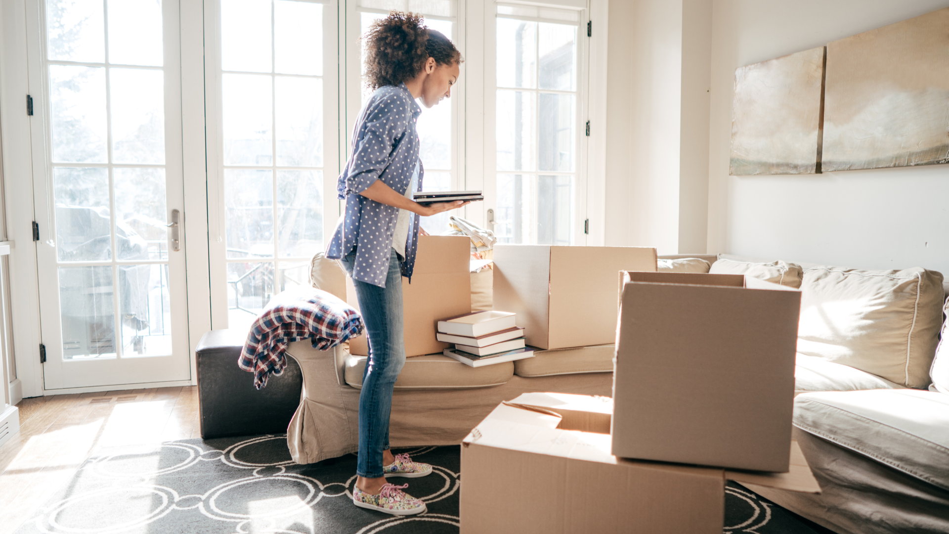 Woman in a room with boxes, looking at a tablet, near windows and a couch.