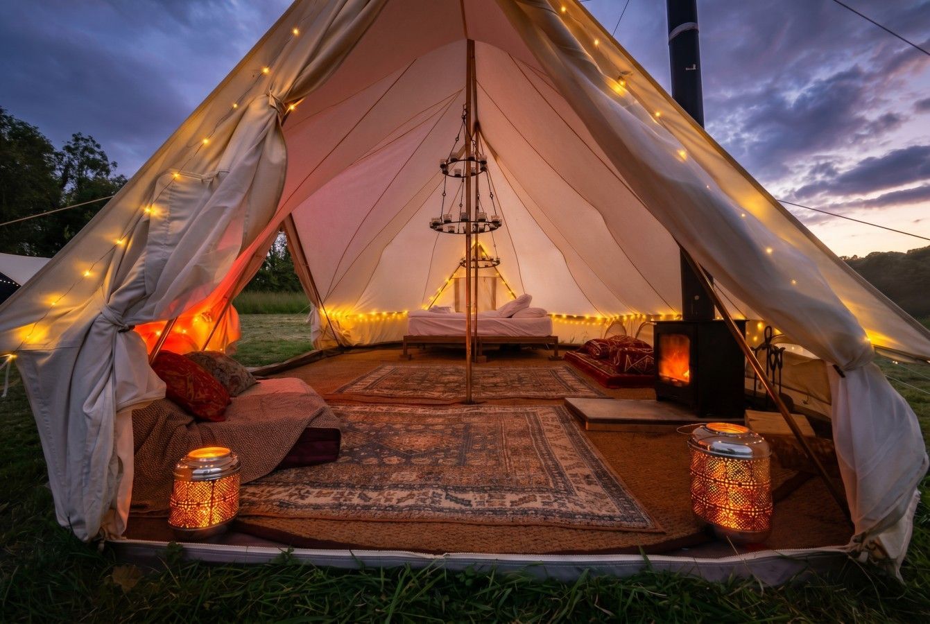 Cozy lit teepee tent interior at dusk, with rugs, lanterns, and warm string lights under a cloudy sky