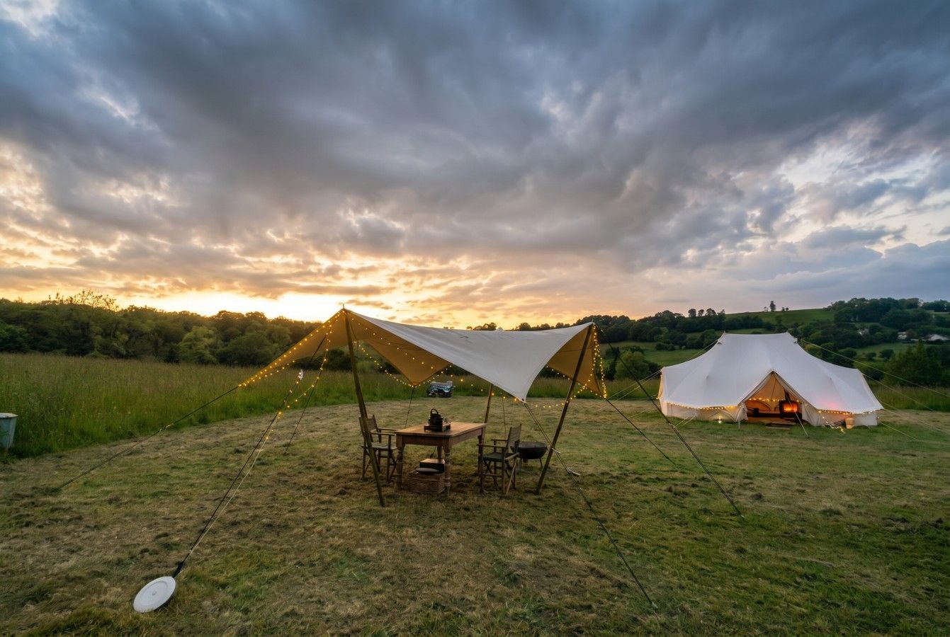 A campsite at sunset with two tents, a tarp shelter, and a fire pit in an open grassy field.