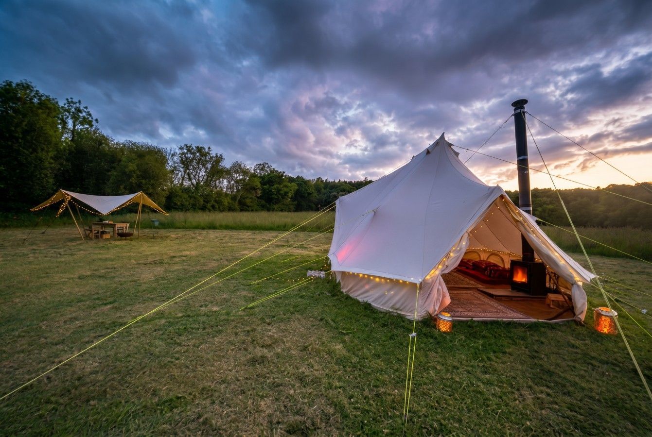 White tent lit at dusk on a grassy field under a dramatic cloudy sky, with another tent in the distance