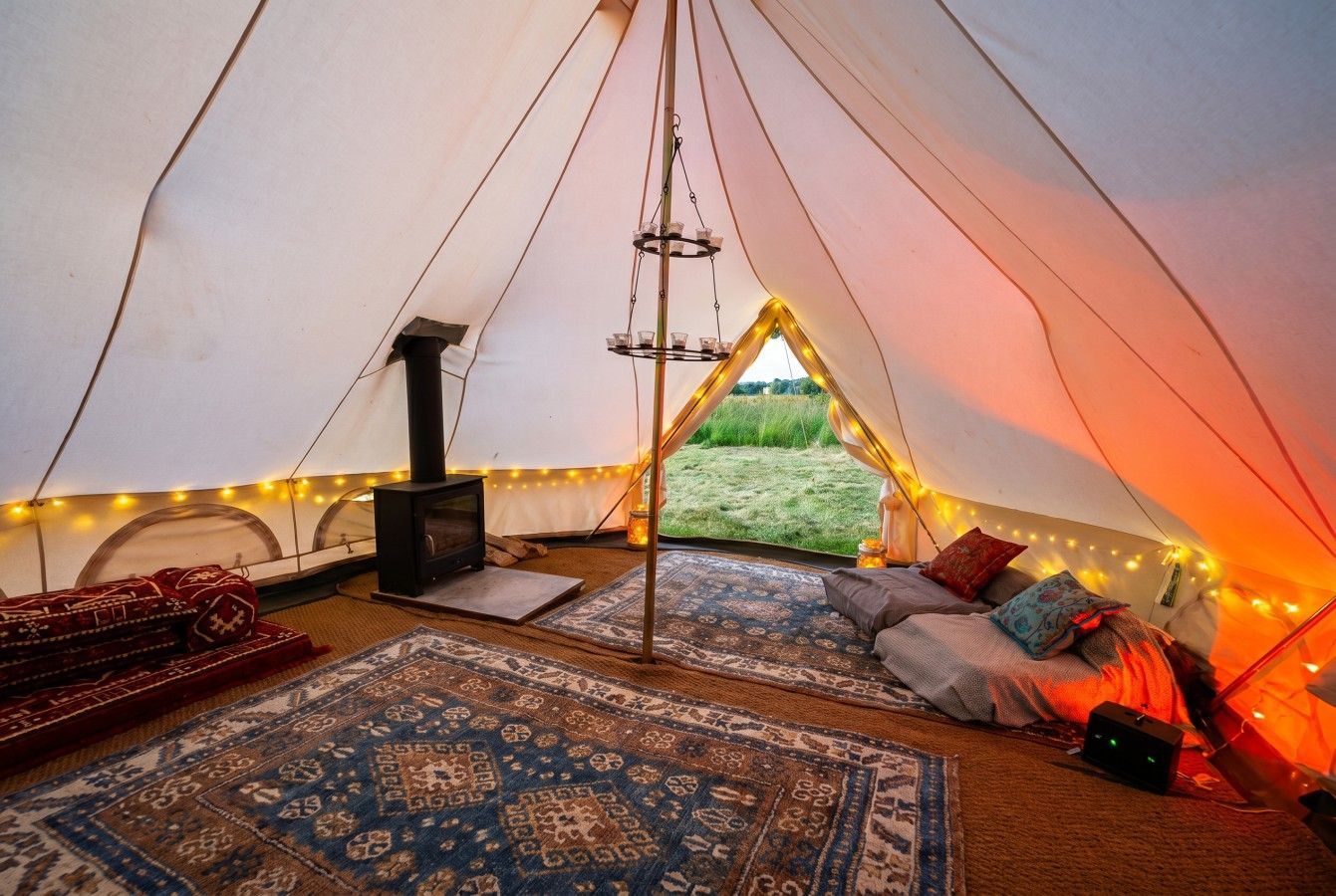 Cozy tent interior with rugs, pillows, wood stove, string lights, and open view to greenery.