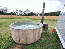 Round wooden hot tub outdoors on grass near a field, with water and a metal stove pipe.