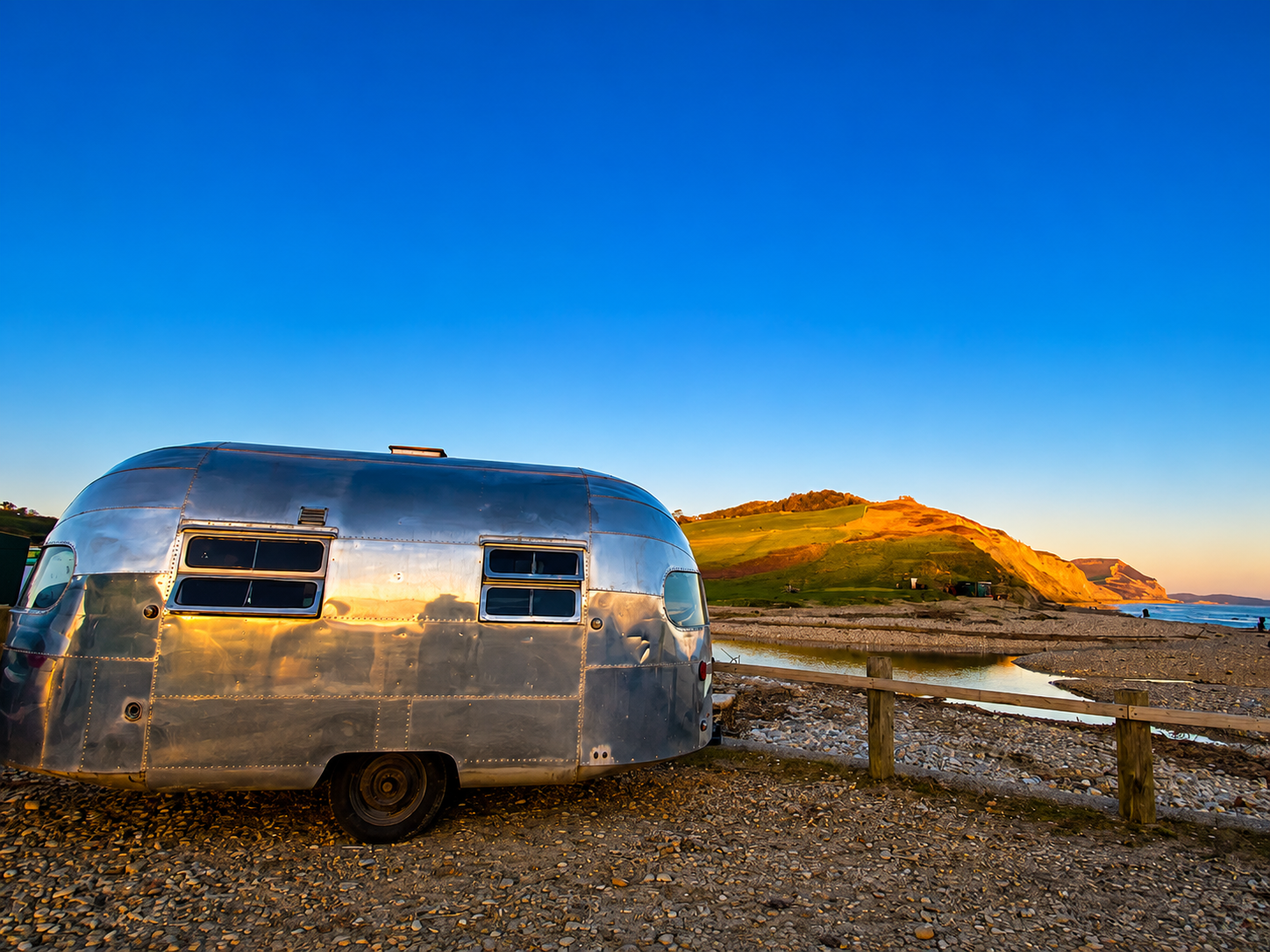 Shiny silver trailer by a seaside fence at sunset, with rocky coast and blue sky.