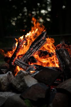 Bright orange flames burning logs in a dark outdoor fire pit, with rocks in the foreground