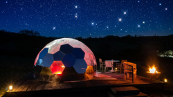 Geodesic dome tent glowing red in a campsite under a starry night sky, with a small fire nearby