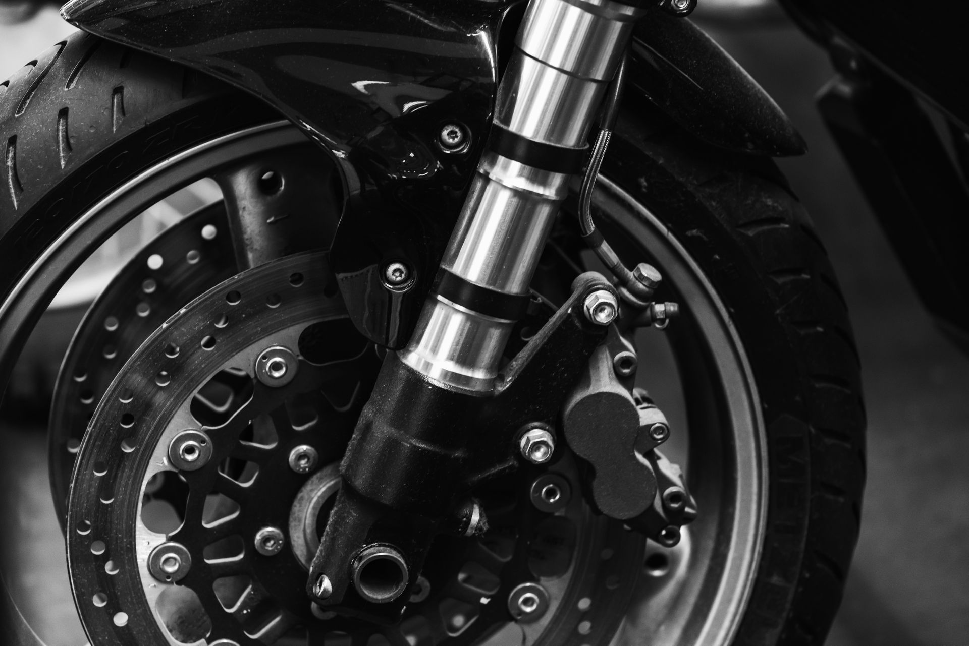 A close-up, black and white shot of a motorcycle's front wheel, brake disc, and suspension fork.