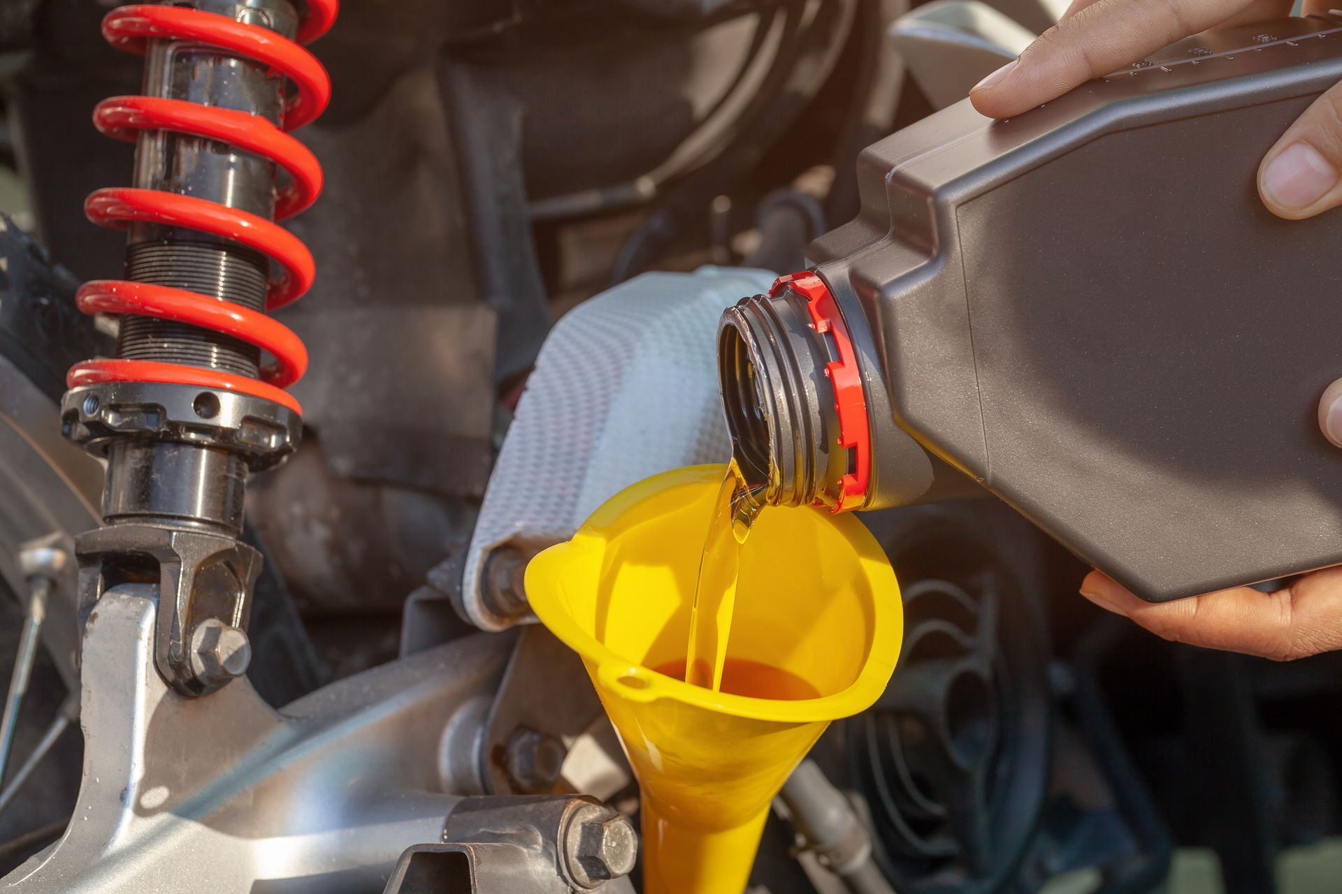 A hand pours golden oil from a bottle into a bright yellow funnel being used to fill a motorcycle's engine.