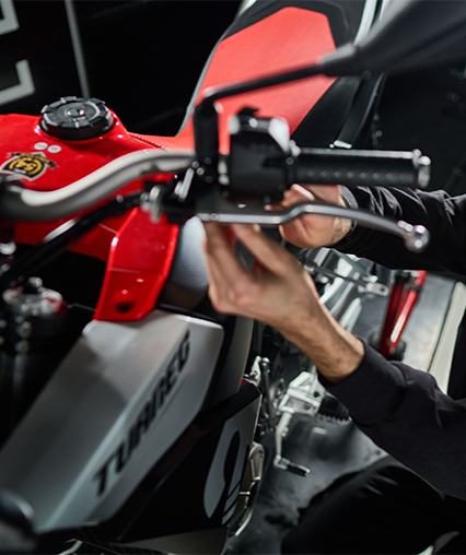 A mechanic in work overalls kneels beside a motorcycle, writing on a clipboard while inspecting the engine.