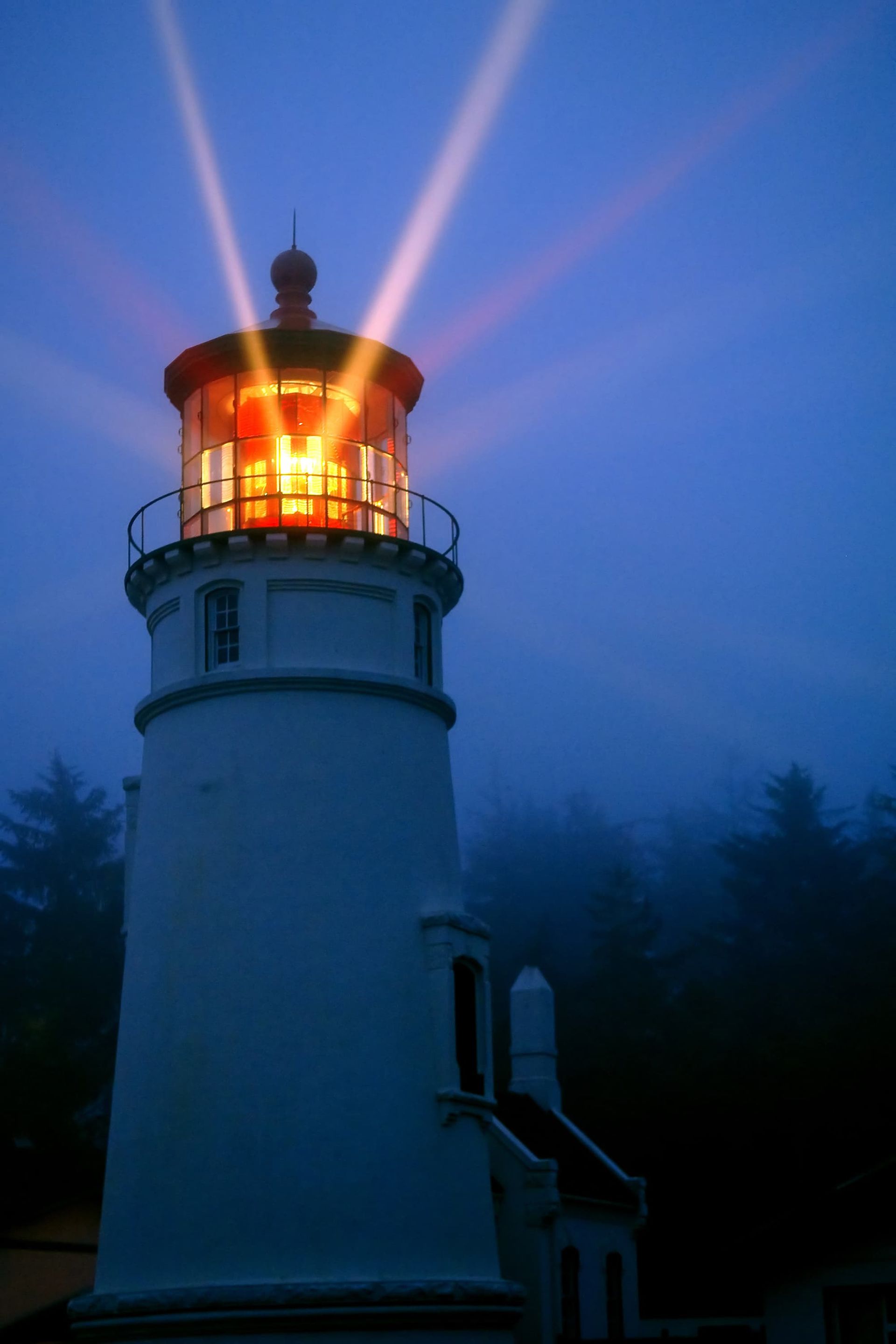 White lighthouse with bright orange light beams cutting through a dark blue, foggy sky.