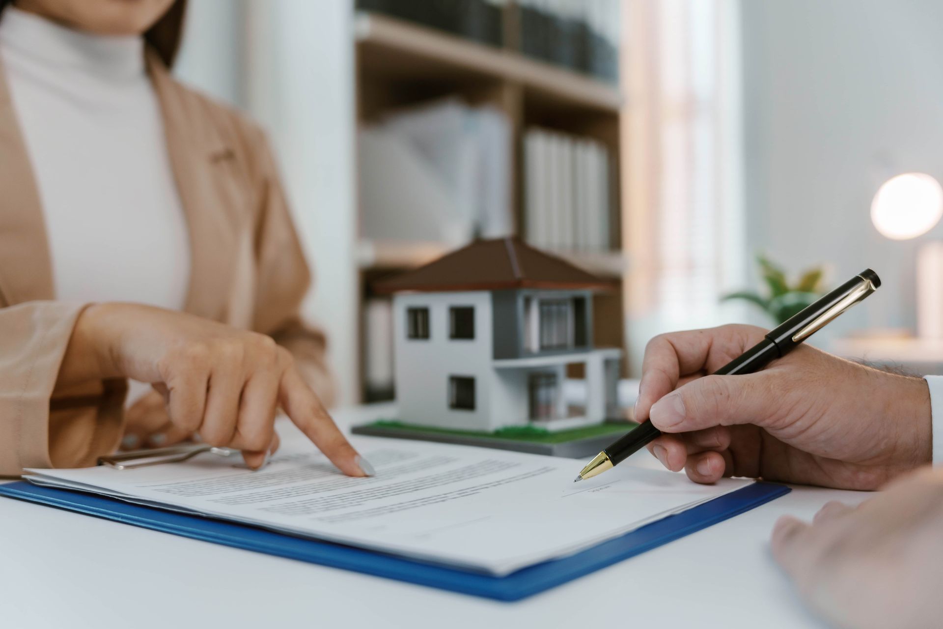 Hands reviewing a document with a pen beside a small house model on a desk