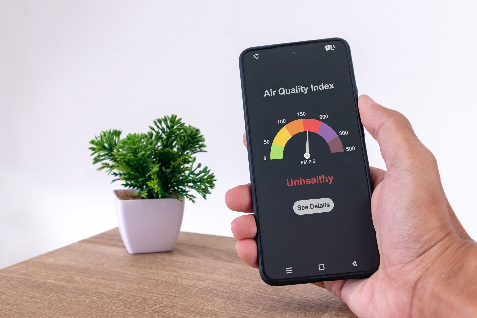 Hand holding smartphone showing air quality gauge near a small potted plant on a wooden table