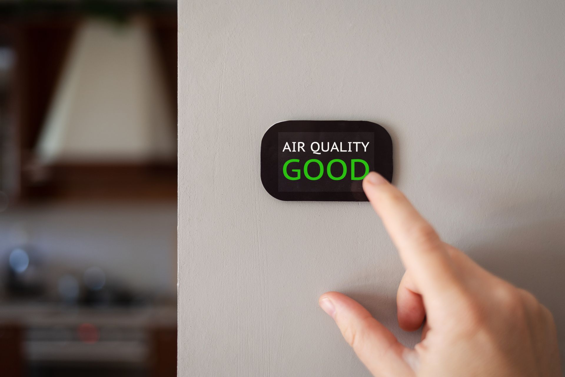 Hand touching a wall-mounted air quality display showing “GOOD” in green.