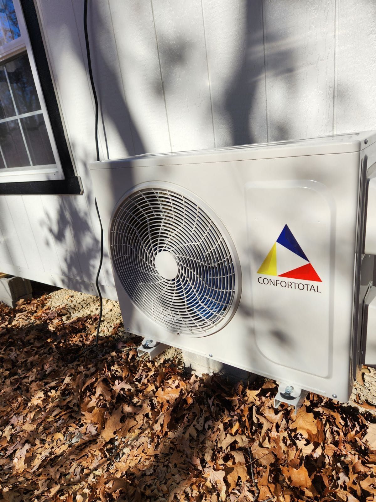 Outdoor HVAC unit on a house wall with a fan, logo, and fallen leaves beneath it