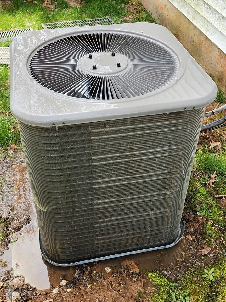 Outdoor air conditioning unit on grass beside a house, with water pooled at the base.