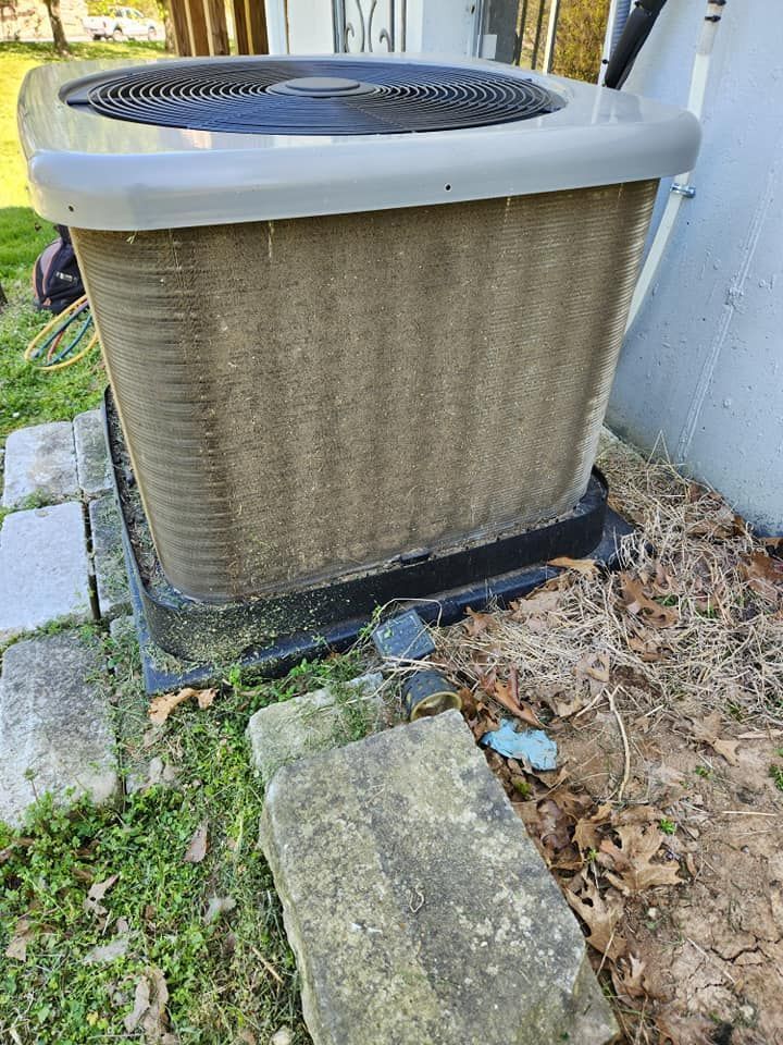 Outdoor HVAC unit beside a wall, with a cracked concrete pad and overgrown grass and leaves nearby