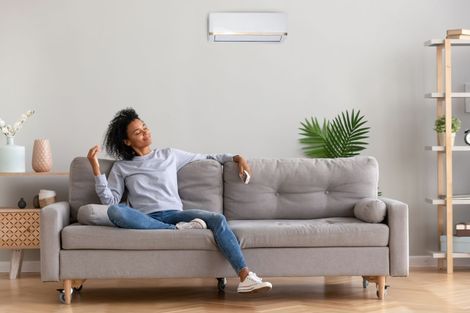 Person relaxing on a gray sofa in a bright living room with a wall-mounted air conditioner and plants