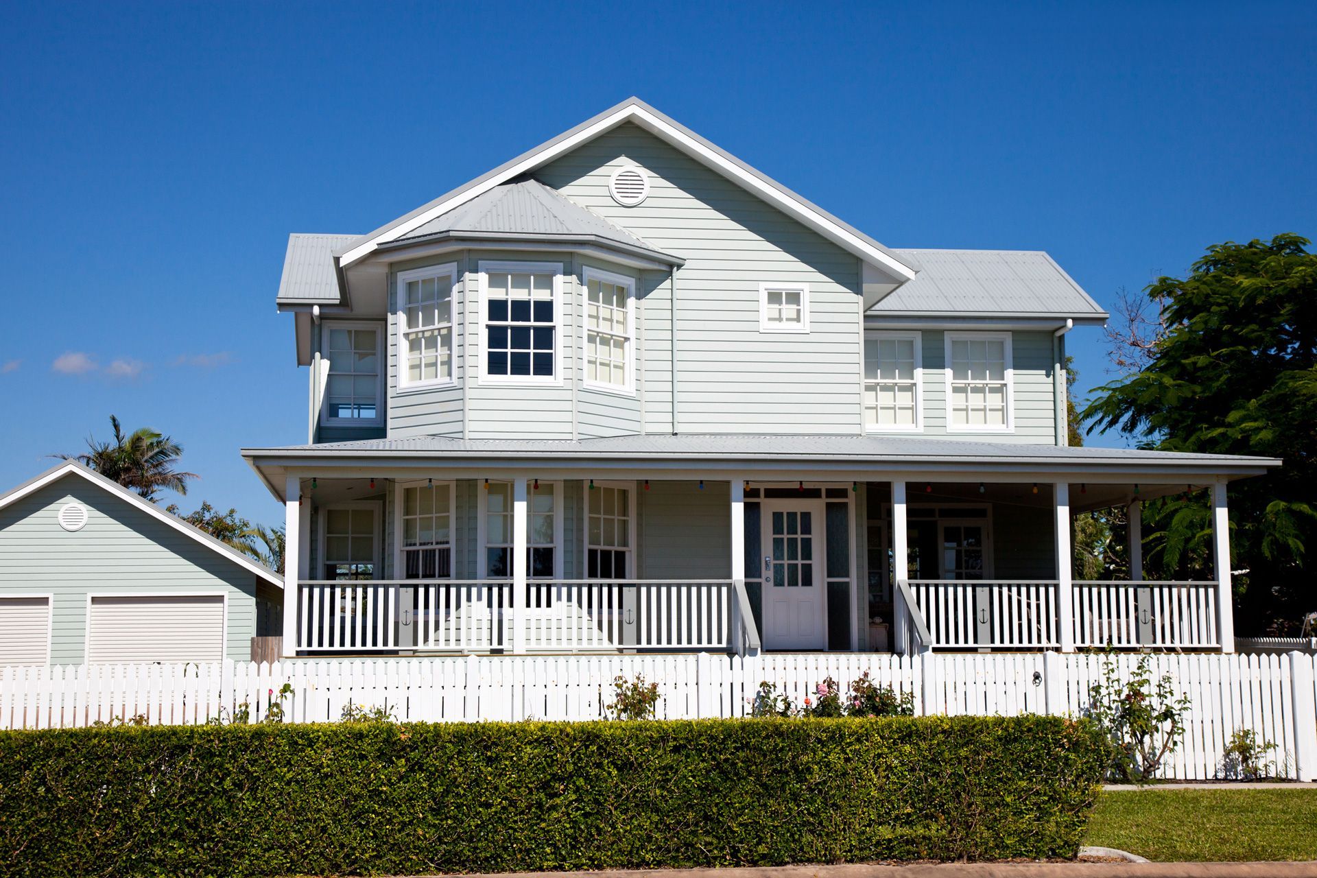 Two-story light blue house with white trim, porch, and picket fence under a blue sky.