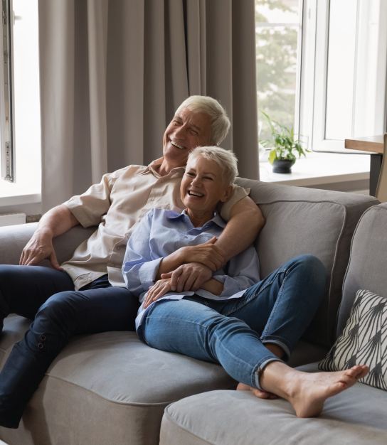 Smiling couple relaxing on a gray sofa in a bright living room, sitting close together by a window.